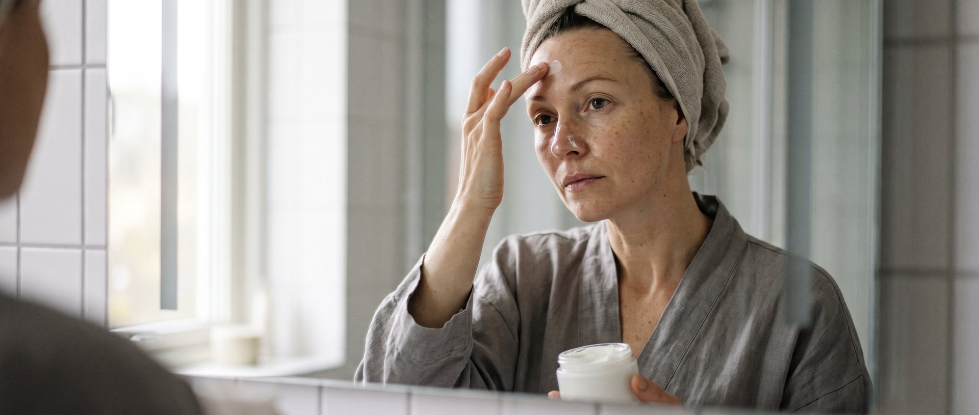 AI-generated illustration of a woman in her forties smoothing moisturizer across her forehead at a bathroom mirror